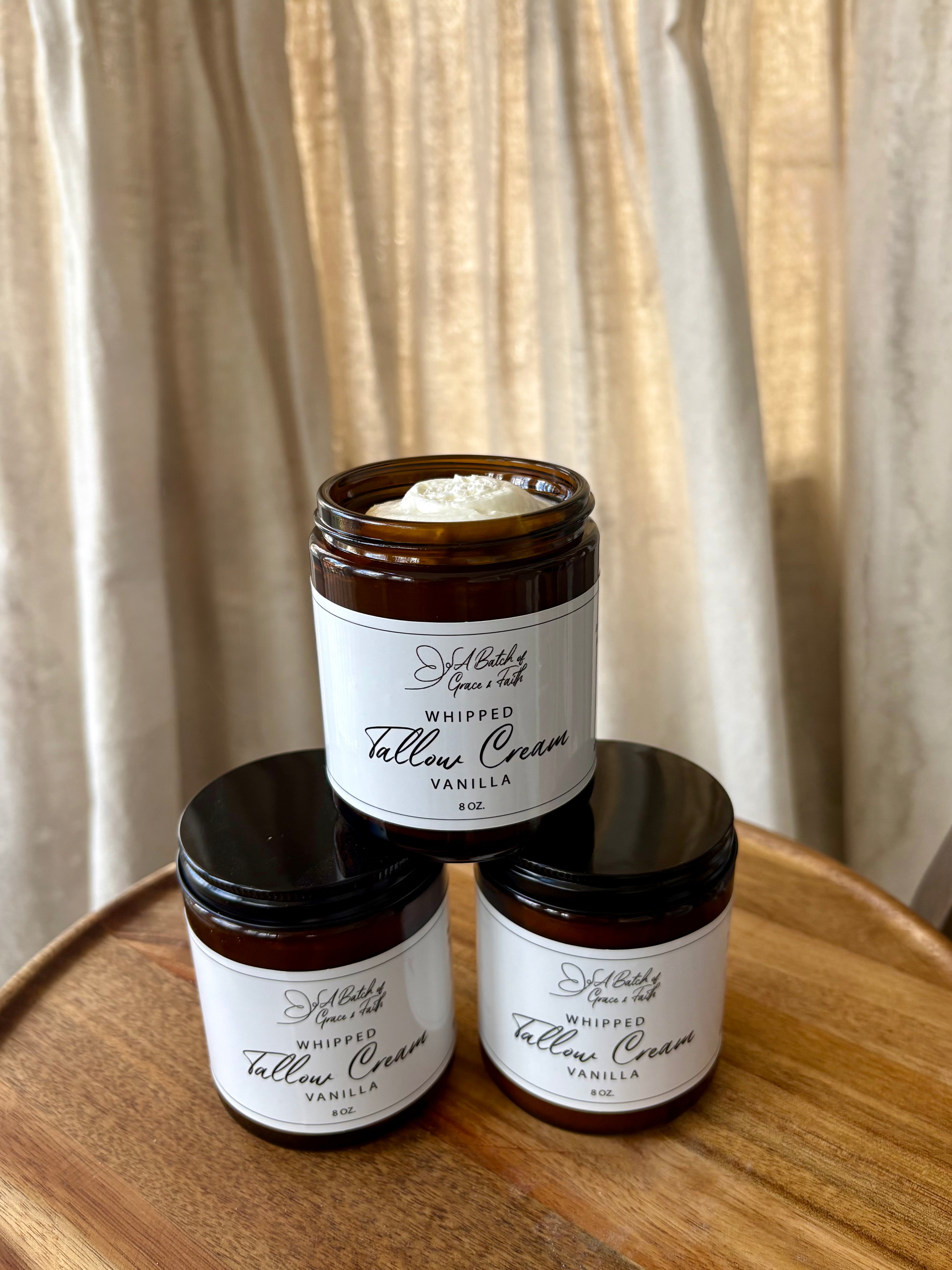 Three jars of 'Whipped Tallow Cream Vanilla' on a wooden surface with a neutral curtain background.